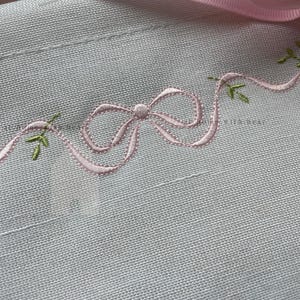 May include: Close-up of a light gray fabric with a delicate pink embroidered bow and trailing ribbon. Small green embroidered leaves accent the ribbon. The fabric has a subtle texture, and the embroidery is finely detailed.