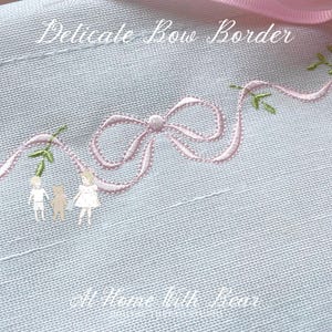 May include: A pale blue fabric featuring a pink embroidered bow and green leaf sprigs. The text "Delicate Bow Border" is above the bow. The text "At Home With Bear" is at the bottom.