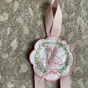 May include: A pink fabric hanger with a white embroidered circle featuring a floral wreath and the letter "H". The hanger has two pink ribbon ties.