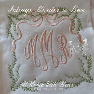 May include: Embroidered design on a white fabric featuring the text "Foliage Border w Bow" and "At Home with Bear". The design includes a pink bow, initials, and a green foliage border. Digital Thread Studio.