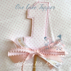 May include: A pink fabric cake topper shaped like the number one, with a stitched outline. It features a pink bow with a ribbon that says "hello little one". The background is pink fabric with small white polka dots. The text "One Cake Topper" is at the top.