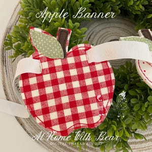 May include: A fabric apple banner with a red and white checkered pattern. The apple has a green polka-dot leaf and a brown stem. White ribbon is attached for hanging. The banner is surrounded by greenery and has the text "Apple Banner" and "At Home With Bear".