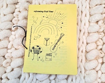 Puede incluir: Un folleto amarillo titulado "reframing lost time" con ilustraciones dibujadas a mano de un arcoíris, un sol, notas musicales y otros símbolos. El folleto está encuadernado con una cuerda negra y descansa sobre una manta de punto grueso color crema.