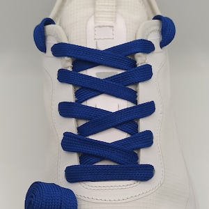 Può includere: Sneakers bianche con lacci blu reale annodati in un motivo a croce. Un rotolo di lacci blu reale si trova nell'angolo in basso a sinistra dell'immagine.