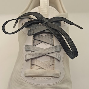 Op de afbeelding: Witte sportschoen met zwarte en grijze veters die in een kruispatroon zijn geknoopt.