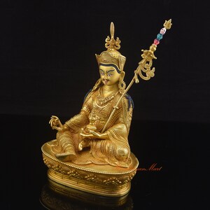 May include: A gold-colored statue of a deity sitting in a meditative pose. The statue is adorned with intricate details and is holding a staff with a skull-shaped finial. The statue is sitting on a lotus throne.