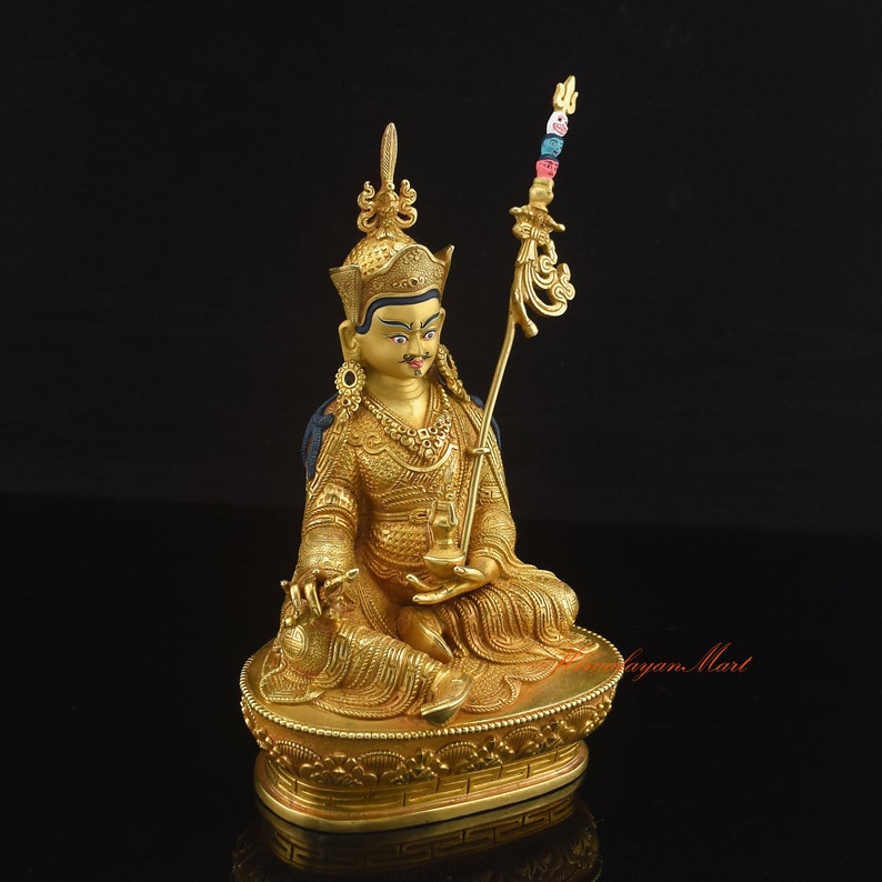 May include: A gold-colored statue of a seated figure with a crown and a scepter. The figure is holding a small object in its right hand. The statue is on a base with a decorative design.