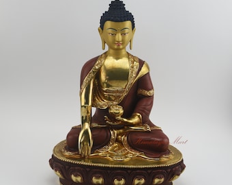 May include: A gold and red Buddha statue sitting in meditation with a lotus flower in his hand. The statue is on a red and gold base with a lotus design.