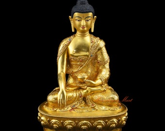 May include: A gold-colored statue of Buddha sitting in a meditative pose. The statue is adorned with intricate details and colorful gemstones. The Buddha is wearing a robe and has a serene expression on his face.