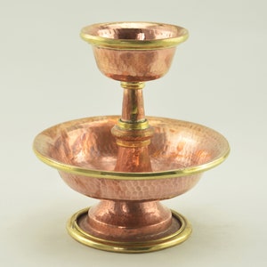 Könnte beinhalten: Eine Wasseropfer Schale aus Kupfer und Messing mit gehämmerter Oberfläche. Die Schale hat einen erhöhten Mittelteil mit einer kleineren Schale oben drauf. Beide Schalen haben einen goldfarbenen Rand.