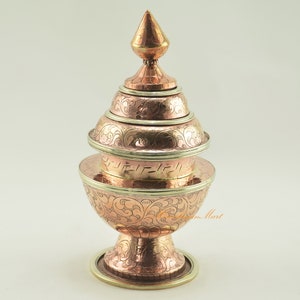 May include: A copper and brass tiered offering bowl with intricate floral engravings. The bowl has a pointed finial on top and a wide base.