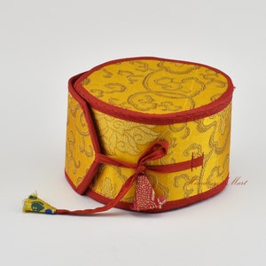 May include: A round, gold-coloured box with a red trim and a red ribbon tied around it. The box is covered in a patterned fabric with a floral design. The box has a tassel hanging from it.