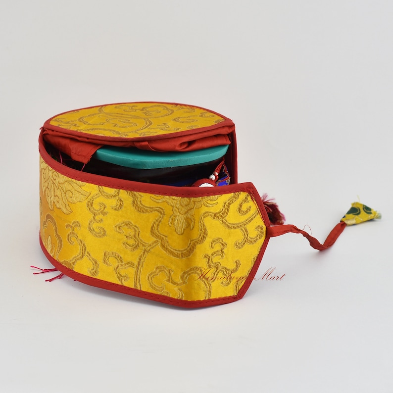 May include: A yellow and red fabric prayer wheel case with a tassel. The case is open and the prayer wheel is visible inside.