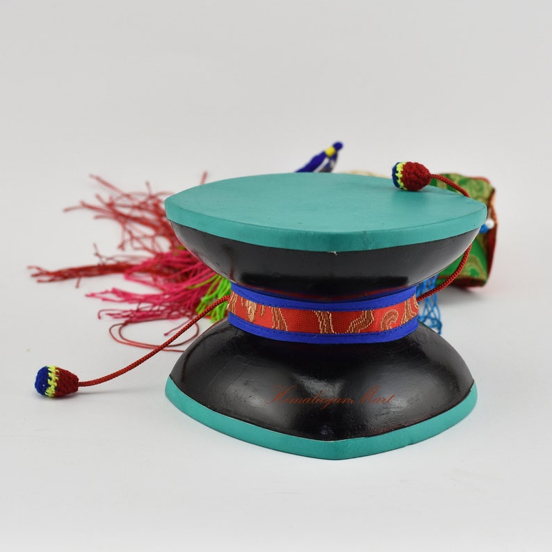 May include: A black prayer wheel with a teal top and red, blue and gold trim. The wheel is attached to a long red cord with a tassel at the end.