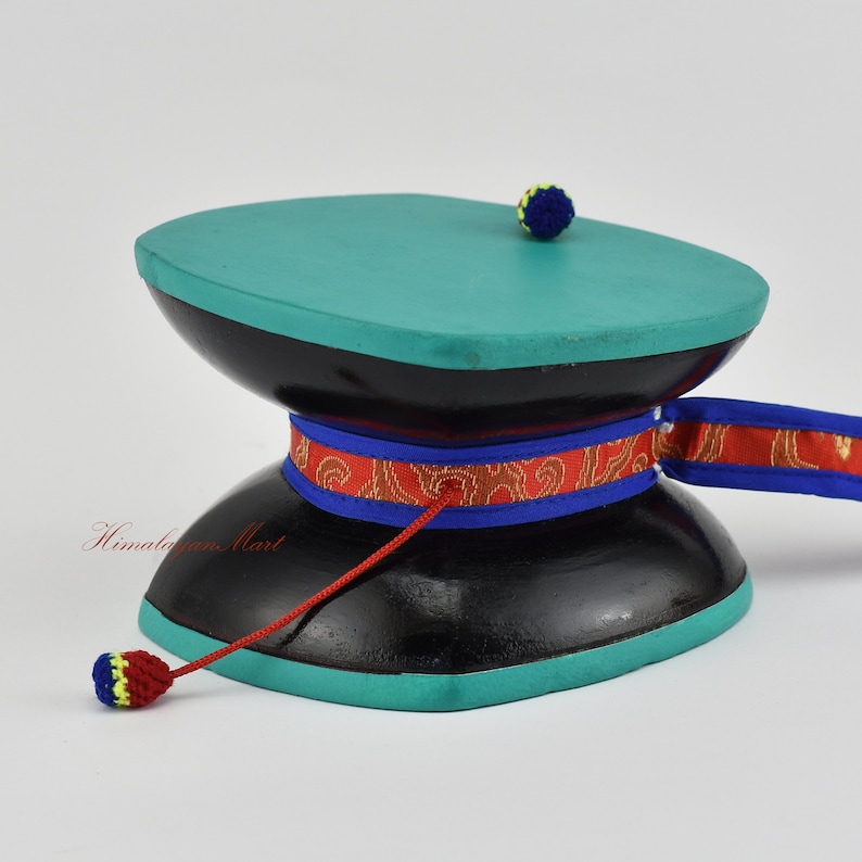 May include: A black prayer wheel with a turquoise top and a red cord with tassels. The wheel is decorated with a red and blue ribbon with gold designs.