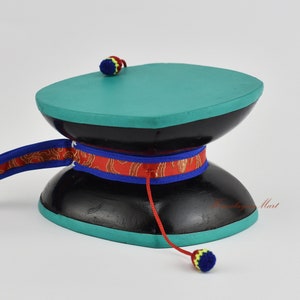 May include: A black and teal wooden prayer wheel with a red, blue and gold ribbon. The wheel has a small red and yellow tassel on the top and a larger blue, yellow and red tassel on the bottom.