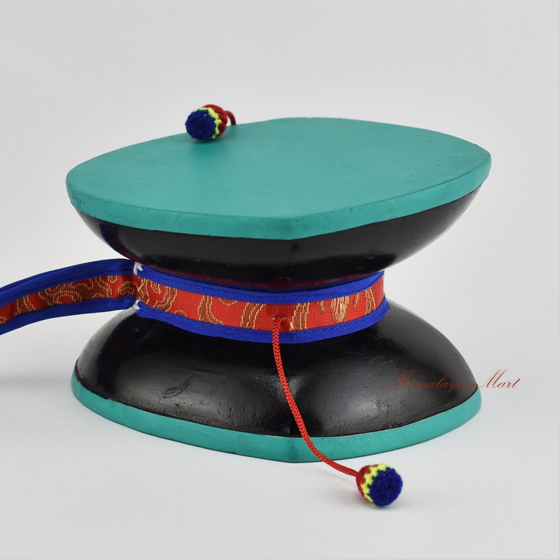 May include: A black and teal wooden prayer wheel with a red, blue and gold ribbon. The wheel has a small red and yellow tassel on the top and a larger blue, yellow and red tassel on the bottom.