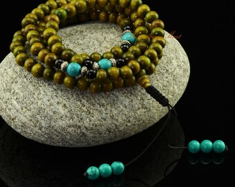 May include: A green wooden mala bead necklace with turquoise and black beads. The necklace is wrapped around a gray stone and has a black cord with turquoise beads at the end.