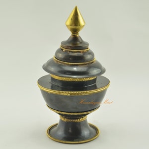 May include: A black metal incense burner with gold accents and a pointed finial. The burner has a round base and a cylindrical body with a lid. The burner is decorated with a gold band and a geometric pattern.