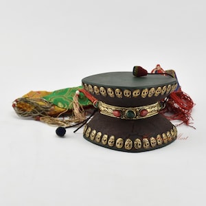 May include: A dark brown wooden prayer wheel with a green top and gold trim. The wheel is decorated with small skull-shaped designs and has a red and green tassel. The wheel is sitting on a colorful fabric.