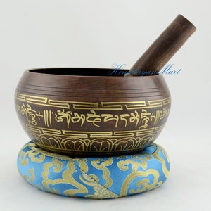 Op de afbeelding: Een bruine zangkom met gouden belettering en ingewikkelde ontwerpen. De kom staat op een blauw en gouden kussen.