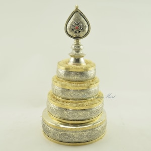 May include: A silver and gold prayer wheel with intricate designs and a red jewel on top. The wheel is made of multiple stacked cylinders with a gold trim.