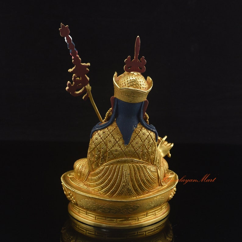 May include: A gold-colored statue of a seated figure with a blue robe and a gold crown. The figure is holding a staff with a red and white flag. The statue is sitting on a gold-colored base with intricate details.