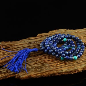 Op de afbeelding: Een blauwe lapis lazuli mala met een blauwe kwast en turquoise accent kralen. De mala ligt op een houten oppervlak.