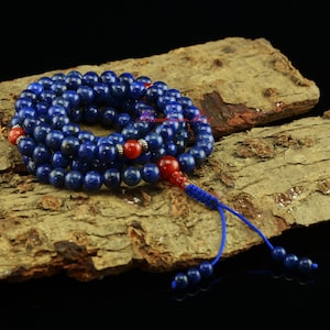 Op de afbeelding: Een streng diepblauwe lapis lazuli kralen met rode accentkralen en een blauwe kwast. De kralen zijn gerangschikt op een houten oppervlak. De kralen zijn ongeveer 8 mm in diameter.