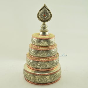Puede incluir: Una rueda de oración de plata con diseños intrincados y una base de cobre. La rueda está coronada con un remate decorativo con una joya roja.
