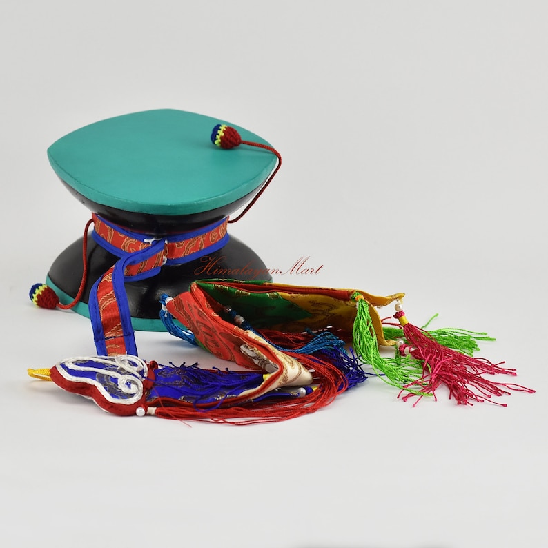 May include: A teal and black prayer wheel with a colourful prayer flag and tassels. The prayer wheel is on a white surface.
