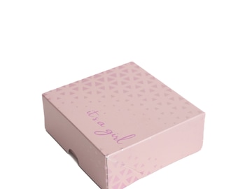 Könnte beinhalten: Eine rosa Geschenkbox mit einem weißen Dreiecksmuster und dem Text "it's a girl" in lila Schrift.