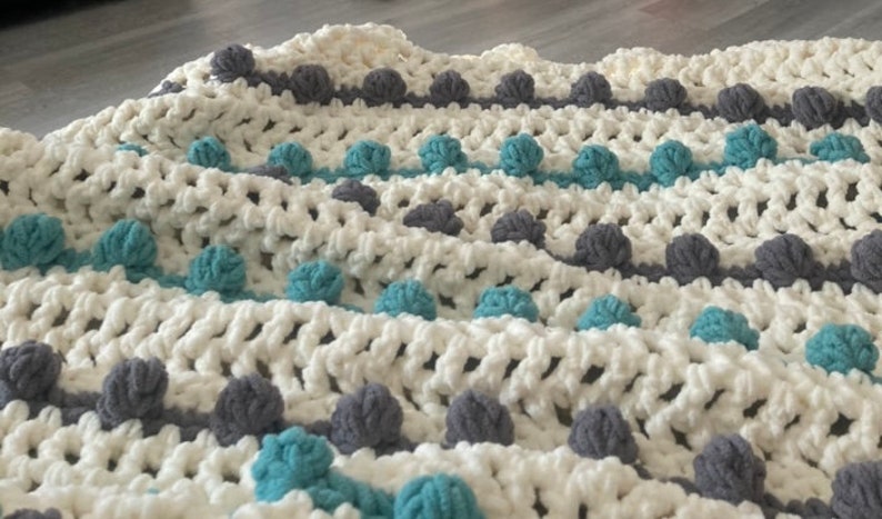 Puede incluir: Manta de crochet blanca con pompones grises y azul turquesa. La manta tiene un patr&oacute;n a rayas con filas alternas de pompones grises y azul turquesa.