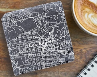 May include: Two black square coasters with a white map of Los Angeles printed on them. The coasters are on a wooden surface with a cup of coffee and a notebook.