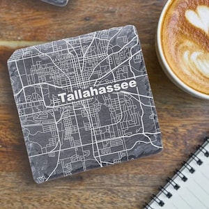 May include: Two gray slate coasters with a white map of Tallahassee, Florida printed on them. The coasters are on a wooden surface with a cup of coffee and a notebook.