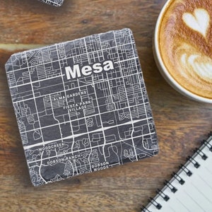 May include: Two black square coasters with a white map of Mesa, Arizona. The coasters feature the city name in white text. The coasters are on a wooden surface with a cup of coffee and a notebook.