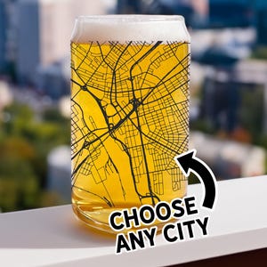 May include: A clear glass beer can-shaped glass filled with beer and foam. The glass features a black and yellow city map design. The text "CHOOSE ANY CITY" is displayed on the glass with a black arrow. The background shows a cityscape.