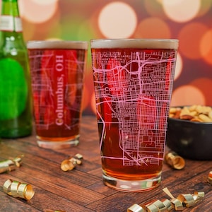 May include: Two clear glass beer mugs with a pink outline of the city of Columbus, Ohio. The text "Columbus, OH" is printed on the side of the mug. The mugs are sitting on a wooden table with gold ribbon.
