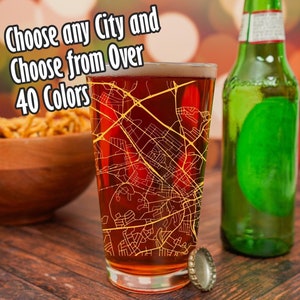 May include: A clear glass beer stein with a city map design printed on it. The map is in yellow and the background is a dark red. The stein is sitting on a wooden table next to a green glass bottle and a bottle cap. The text "Choose any City and Choose from Over 40 Colors" is visible in the image.