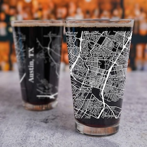 May include: Two clear glass pint glasses with black liquid inside. The glasses feature a white outline of a city map with the text "Austin, TX" printed on the side.