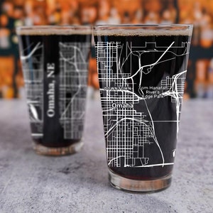 May include: Two pint glasses filled with a dark liquid, possibly beer, are displayed. Each glass features a white map design of Omaha, NE, with street names and landmarks. The glasses are on a gray surface.