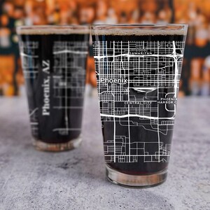 May include: Two clear glass beer mugs with black liquid inside. The mugs have a white outline of the city of Phoenix, Arizona, printed on them. The text "Phoenix, AZ" is visible on the mug in the background. The text "Phoenix" and "Central City" are visible on the mug in the foreground.