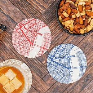 May include: Two round coasters with a map of New Orleans printed on them. The coasters are white with red and blue lines representing streets. The coasters are on a wooden surface with a bowl of mixed nuts in the background.