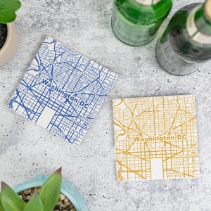 May include: Two square coasters with a map of Washington DC. The top coaster has a blue map with white lines and the bottom coaster has a yellow map with white lines. Both coasters have the text "Washington DC" on them.