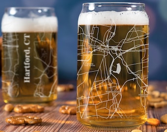 May include: Two clear glass beer mugs with a map of Hartford, CT etched on the side. The mugs are filled with a golden-colored beer. The mugs are sitting on a wooden table with pretzels scattered around them.