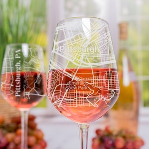 May include: Two clear wine glasses with a map of Pittsburgh, Pennsylvania etched on them. The glasses are filled with rose wine and are sitting on a table with grapes in the background.