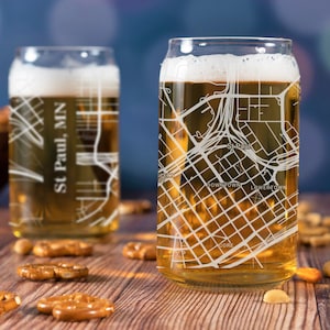 May include: Two clear glass beer can glasses with a white outline of the city of St. Paul, Minnesota etched on the side. The glasses are filled with a light golden beer and are sitting on a wooden table with pretzels and nuts.