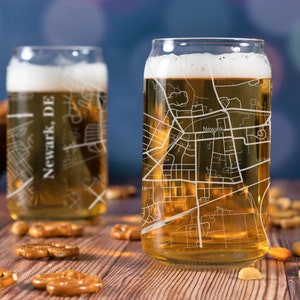 May include: Two clear glass beer mugs with a city map design. The mug on the right has the text "Newark" on the map. The mugs are filled with a light golden beer and are sitting on a wooden table with pretzels and nuts.