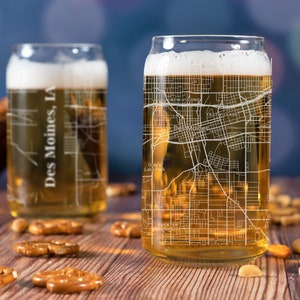 May include: Two clear glass beer mugs with a white outline of the Des Moines, IA city map. The mugs are filled with a golden yellow beer and are sitting on a wooden table with pretzels and nuts.