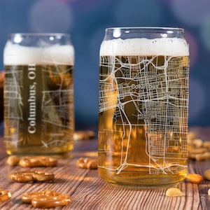 May include: Two clear glass beer mugs with a white outline of the city of Columbus, Ohio. The mugs are filled with a light golden beer and are sitting on a wooden table with pretzels and nuts.